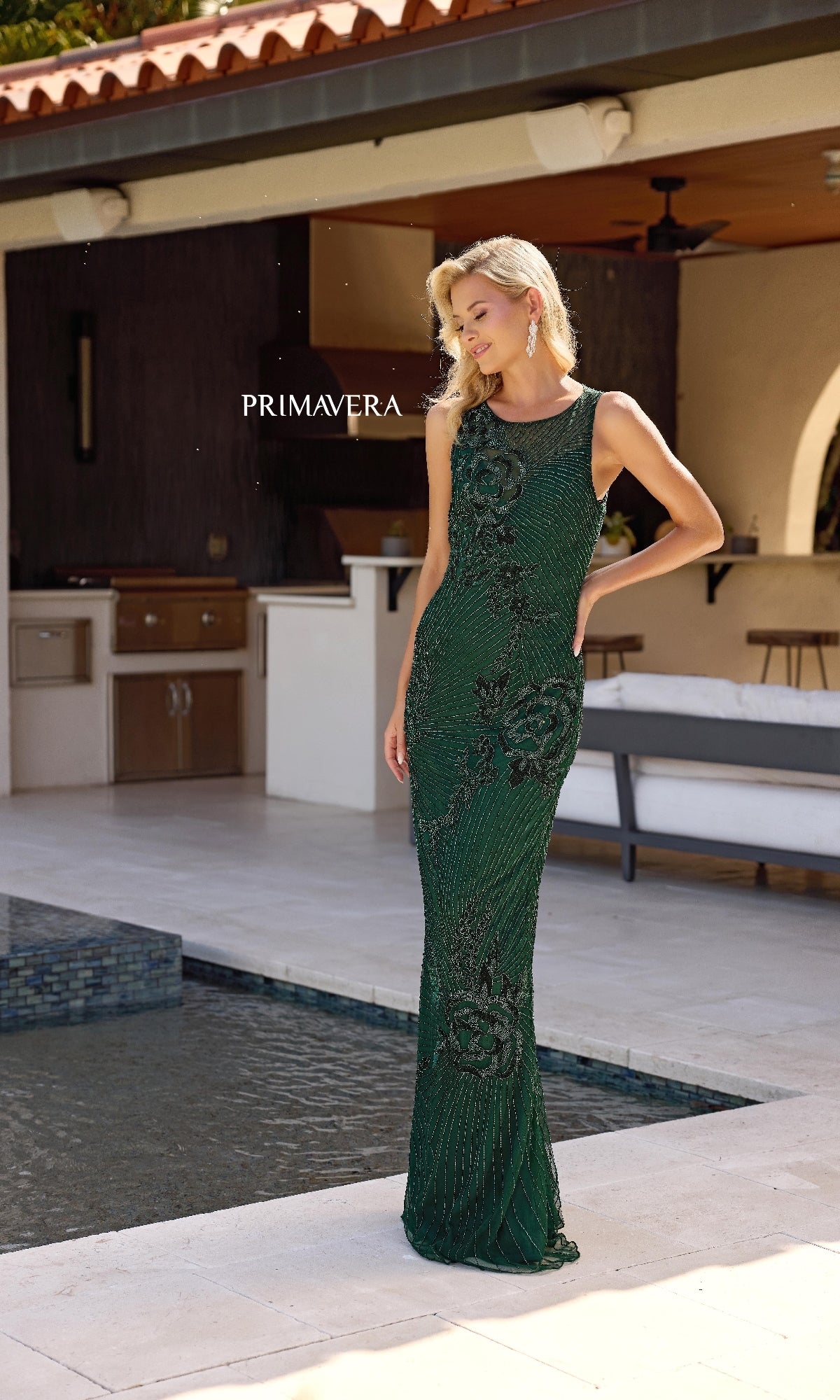 A woman in the Primavera 13140 sleeveless long formal dress with an illusion-sweetheart neckline poses and smiles slightly by a modern outdoor pool area.
